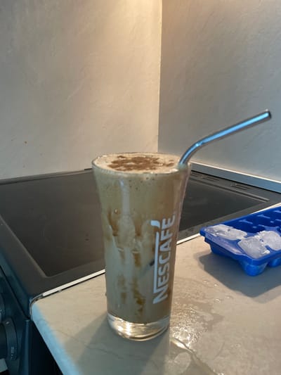 In Zlatograd, Bulgaria, a glass of iced coffee with a Nescafé logo and a metal straw sits on a kitchen counter. The drink is topped with a dusting of cinnamon. To the right of the glass, a blue ice cube tray holds several ice cubes. The counter surface is slightly wet around the glass. In the background, a stovetop and a tiled wall are visible.