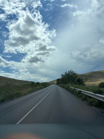 The image depicts a view from a vehicle traveling along a two-lane asphalt road. The road curves slightly to the right, with a white dashed line marking the center. To the left of the road, a grassy hillside slopes upwards, dotted with sparse vegetation. To the right, a metal guardrail separates the road from a dense growth of green bushes and trees. Beyond the guardrail, the terrain consists of a gently sloping, dry, yellowish-brown hillside. In the distance, several trees are visible on the right side of the road, and two white vehicles appear to be on the road ahead. The sky is filled with dramatic, textured clouds, ranging from bright white to darker grays, against a backdrop of blue. The overall impression is that of a scenic drive through a rural landscape under a dynamic sky, possibly in the vicinity of Isperihovo, Bulgaria.