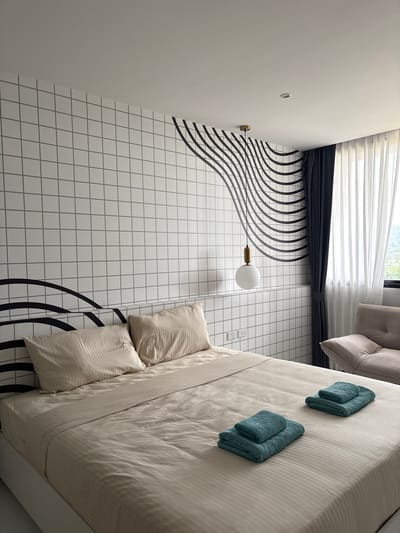 A modern bedroom in Rawai, Thailand, features a queen-sized bed with beige, striped bedding and two light beige pillows. Two stacks of teal towels are placed on the bed. The wall behind the bed is covered in white square tiles with grey grout, decorated with a black wavy line pattern. A white pendant light with a brass accent hangs from the ceiling above the bed. To the right, dark gray curtains are drawn partially, revealing sheer white curtains and a window. A light brown recliner is visible through the curtains. The room appears to be a hotel room or a similar accommodation.