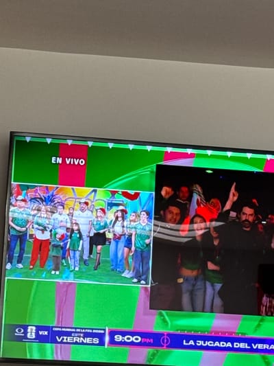 The image is a television screen displaying a live broadcast. The screen is divided into two main sections. The left side shows a group of people, including men, women, and children, standing on a stage with a green and pink backdrop. Many of the individuals are wearing green and white attire, resembling sports fan clothing. The text "EN VIVO" (meaning "live" in Spanish) is prominently displayed at the top of this section.

The right side of the screen is darker and shows a crowd of people with some individuals raising their arms, suggesting an enthusiastic audience.

The bottom portion of the screen contains broadcast information. The text "COPA MUNDIAL DE LA FIFA 2020 ESTE VIERNES 9:00PM LA JUGADA DEL VERANO" is visible. This translates to "FIFA World Cup 2020 This Friday 9:00 PM The Play of the Summer." The presence of "VIX" suggests a media platform or channel.

The image is a captured photograph of the television screen itself, and the television is situated in a room with a plain wall. There is no visual information within the image that allows for the determination of a specific location in San Bernardino Tlaxcalancingo, Mexico.