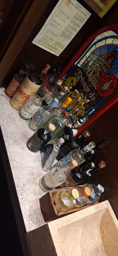 The image captures an eclectic bar setting, likely in Venice, Italy, considering the prevalence of Italian text on a document. The scene is dominated by a collection of liquor bottles arranged on a patterned surface, indicative of a countertop or bar top.
Among the bottles, brands such as "Ron Papa Rum," "Gin Sospiri," "Kino," "Caprisius Gin," and "Amaro Ostrano" are visible, suggesting a selection of rum and various gins. The arrangement appears somewhat disorganized, implying either a recent service or an aesthetic choice.
Above the bottles is a stained-glass style decoration featuring the words "Abbaye de," hinting at a possible thematic element in the bar's decor or a reference to a particular spirit or drink.
To the left, a document titled "ELENCO" (Italian for "list") is present, containing what appears to be a menu or list of offerings in Italian.
Adjacent to the bottles on the right is a stone-like structure, perhaps a sink or part of the bar's design, adding a textural contrast to the scene.
The lighting is dim, creating a cozy atmosphere, with the focus primarily on the bottles and the decorative elements around them. Overall, the image portrays a unique, possibly themed, bar environment in Venice, Italy.