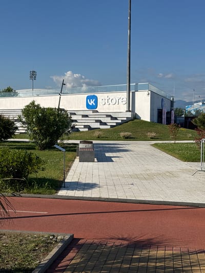 The image shows an outdoor scene at eye level, with a clear blue sky and scattered clouds. A modern building with large glass windows and white facade is visible on the left. A blue logo with a stylized "K" is prominently displayed on the building, along with the word "store" in large white lettering. In front of the building, a series of wide, light-colored stone steps lead up to the entrance. Adjacent to the steps, a grassy slope is covered with neatly mowed grass and some small bushes. A dark gray trash receptacle is positioned on a paved area near the grass.
To the right of the paved area is a red running track with a white line marking its edge. The track is bordered by vegetation, including green bushes and grass, and a concrete curb. Shadows from unseen objects, possibly trees or structures, are cast diagonally across the paved area and the running track, suggesting it is daytime. In the background, faint outlines of distant mountains or hills can be seen. A tall light pole with multiple lamps stands in the distance, and a small bird is perched on a thin pole in the sky. The overall atmosphere is bright and sunny, suggesting fair weather. The scene appears to be a public space, possibly part of a sports facility or commercial complex.
