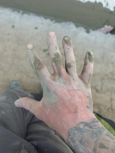 A top-down, eye-level shot reveals a light-skinned human hand covered with a gray, particulate substance. The hand has a tattoo on the wrist, the design of which is not fully discernable. Gray residue is concentrated on the fingers, joints, and the back of the hand, highlighting the texture of the skin. The hand is positioned over a surface with gray pants and a partial view of a gray shoe in the lower right. The surface to the left shows evidence of gray mortar or concrete, potentially a freshly laid surface. The location, Crauthem, Luxembourg, suggests this could be a construction or renovation site, but without further context, this can only be inferred.