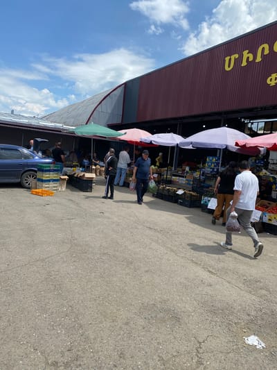 The image captures a bustling outdoor market scene in Gyumri, Armenia, under a bright, partly cloudy sky. In the foreground, a paved ground shows signs of wear and tear. People are seen browsing and interacting with vendors. Several stalls are set up, covered by a variety of colorful umbrellas, including green, pink, and white ones. These stalls display an abundance of fresh produce, with crates of fruits and vegetables visible. A blue car is parked near one of the stalls, with its trunk open, suggesting active trade. The background features a large building with a distinctive burgundy-colored facade, marked with yellow Armenian lettering. The overall atmosphere is that of a lively marketplace where goods are being exchanged.