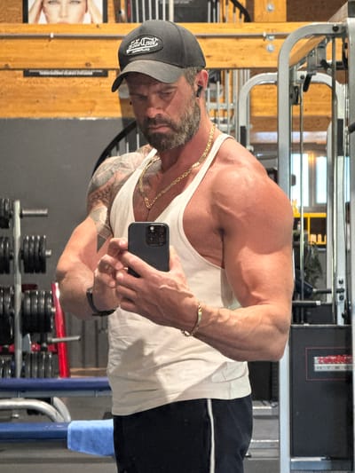 A muscular man with a beard, wearing a gray baseball cap, a white tank top, black shorts, and gold jewelry, is taking a selfie in a gym. The gym equipment and weights are visible in the background. The photo appears to be taken in Jemelle, Belgium, as that is where the images are located.