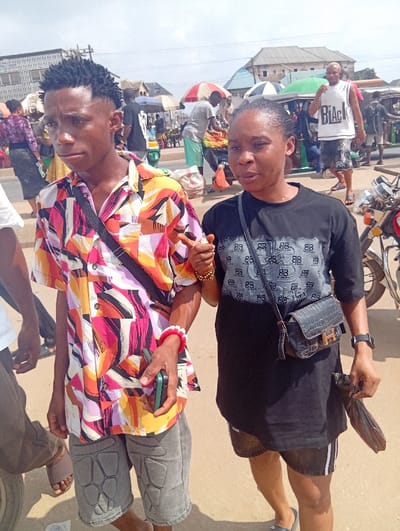 The image depicts a bustling outdoor street scene in Ura Nta Umuarandu, Nigeria. In the foreground, two individuals stand side-by-side. On the left is a young adult male with dark skin, short black dreadlocks styled upwards with faded sides. He wears a short-sleeved collared shirt with a vibrant abstract pattern in pink, orange, yellow, white, black, and purple. He has grey shorts with dark grey padded knee areas, a black crossbody bag strap, a gold-colored bracelet on his left wrist, and a red and white beaded bracelet on his right wrist. He holds a green flip phone in his right hand. To his right stands a young adult female with dark skin and dark hair pulled back. She wears a black short-sleeved t-shirt with a repeated grey "BB" pattern on the chest, dark denim shorts with white stripes, a black crossbody bag featuring a gold "H" emblem, and a black wristwatch on her left wrist. She holds a small black plastic bag in her left hand, and her mouth is slightly open. Both individuals are oriented slightly away from the camera.
The background reveals a busy market environment. Numerous pedestrians are visible, with vendors operating market stalls under colorful umbrellas, including red, white, and green ones. Produce and goods are displayed, and several sacks are present on the ground. A man in a white sleeveless shirt with "BLACK" printed on it and patterned shorts walks towards the background on the right. Parts of motorcycles are visible on the far right. Light-colored buildings with dark roofs are situated in the mid-distance. The ground surface is unpaved.