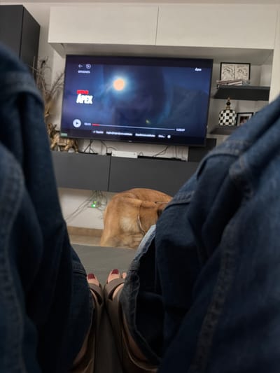 The image shows a first-person perspective of a person sitting on a sofa, wearing blue jeans and beige sandals with red nail polish. A dog is seated near the person. A television is displaying "Apex" through Netflix. The background includes a white cabinet with decorative objects. The location is presumably a living room in Santa Clara Ocoyucan, Mexico, but there's no information that directly confirms this.