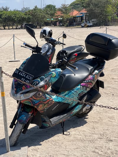 The image depicts two scooters parked on a sandy area, likely a beach or parking lot, in Denpasar, Indonesia. The scooter in the foreground has a license plate that reads "DK 4454 ACH" and is adorned with a vibrant, abstract graphic design featuring swirling patterns and stylized faces. It also has a black windscreen and a black storage box mounted on the rear. A white and black helmet rests on the handlebars of the scooter behind it, which appears to be black and red with the word "sporty" printed on its side. The background shows a line of trees and buildings with orange roofs, suggesting a coastal town or resort area. The scooters are partially enclosed by a chain-link fence. The lighting indicates it is daytime, with shadows cast on the sand.