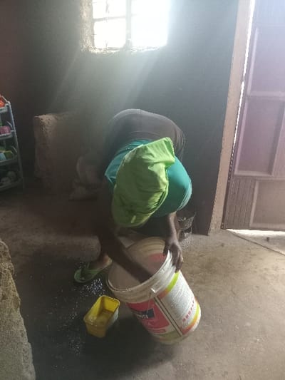 An indoor scene in Manda-Shivanga ward, Kenya, depicts an individual bent forward, actively engaged with a large white bucket. The person wears a dark upper garment, a light green fabric over their back/shoulders, and light green flip-flops on their feet. Their hands are submerged inside the white bucket, which features a multi-colored label including the word "APEXA." Adjacent to the bucket on the dark, damp floor is a yellow rectangular container holding a clear liquid. Splashed liquid is visible on the floor around these items. A bright, gridded window high on the rear wall illuminates the space with strong natural light, casting visible rays through the otherwise dimly lit room. The walls are dark and textured. To the far left, a light-colored, multi-tiered shelving unit holds various small, colorful plastic items. A light-colored door with vertical paneling is partially visible on the right side of the frame. The floor appears to be concrete or compacted earth.