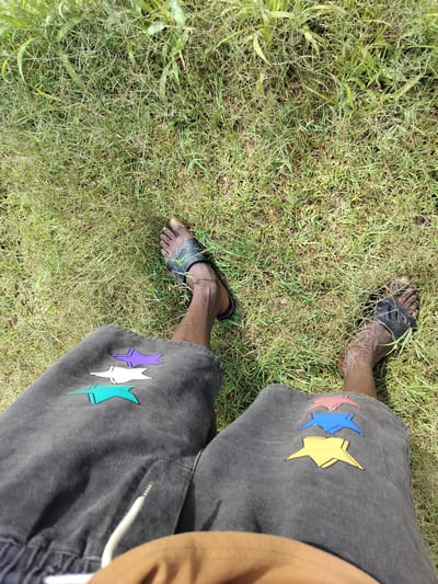 The image shows a person's legs and feet from a high-angle perspective, sitting on a grassy area. The person is wearing dark-colored shorts with colorful star appliques and black slide sandals. The grass is green and somewhat sparse in some areas. There is no indication of the city of Du, Nigeria, or any specific urban environment.