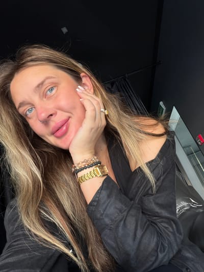 A woman is taking a selfie indoors. She is wearing a black top with her left shoulder exposed and has long, blonde, wavy hair. She is wearing several bracelets on her left wrist, including a gold watch, and a ring with a large yellow stone on her right ring finger. Her left hand is resting on her cheek. The background appears to be dark, with black curtains visible. There is a reflection in a mirror on the right side of the image, showing a green sign with Cyrillic text.