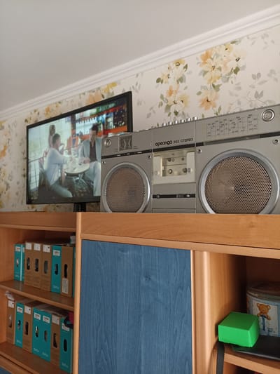 This media file captures an indoor scene, likely a living room or study, with a retro aesthetic. The primary objects are a television and a silver boombox, both positioned on top of a wooden cabinet. The television displays a scene with two people, appearing to be engaged in conversation at a cafe. The boombox, labeled "Ореанда 203 СТЕРЕО," is a prominent object with visible controls and speakers.
The cabinet below the television and boombox contains several shelves filled with stacked boxes. These boxes are primarily brown cardboard with blue labels, some featuring the text "SunLu" and "SUNLU" repeated, suggesting they might contain products from this brand. One shelf also holds a blue decorative cabinet door or panel. To the right, on a shelf, a green cube rests on a black surface, next to a tin box decorated with a teddy bear.
The wallpaper in the background features a floral pattern with pale yellow and cream-colored flowers and green leaves. The lighting suggests it is daytime, with light coming from above and slightly to the left, illuminating the scene and casting soft shadows. The overall atmosphere appears calm and domestic, hinting at a relaxed or leisurely moment. The visible text on the boombox and the boxes provides brand and model information. The location context of Kharkiv, Ukraine, places this domestic scene within that geographical area.
