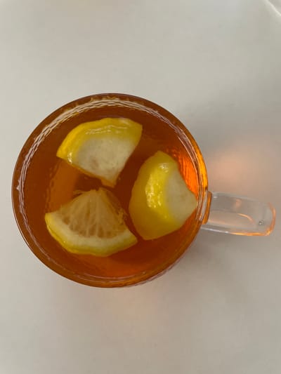 A top-down, medium shot captures a clear glass mug filled with amber-colored liquid and three lemon slices. The mug has a ribbed texture along its rim and a transparent handle extending to the right. The liquid is a rich, warm hue, suggesting tea or a similar beverage. The lemon slices, with their white pith and yellow rind, are partially submerged. The background is a plain, light-colored surface, possibly a table or countertop. The scene appears to be indoors, with soft lighting. There are no people or animals visible. The location in Vologda, Russia, is not visually indicated in the image.