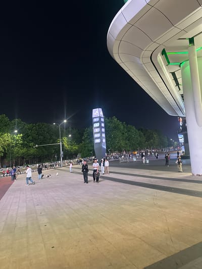 The image depicts an outdoor scene at night, likely in a public urban area. In the foreground, a paved walkway with a geometric pattern is visible, leading towards the right side of the frame. The walkway appears to be made of large rectangular tiles.

To the right, a modern architectural structure with a curved, white facade dominates the upper portion of the image. This structure features integrated green lighting, highlighting its sleek design. White cylindrical pillars support the overhang.

In the mid-ground, a diverse group of people are walking on the paved area. Some individuals are dressed casually, while others are in more formal attire. A tall, illuminated digital display tower with various logos and text is prominently situated to the left of the people. Trees line the background, forming a dark green canopy against the night sky. Streetlights with visible beams illuminate the area, casting a warm glow.

Further in the background, parked motorcycles and scooters are visible, suggesting a transportation hub or a busy area. Fences and barriers are also present, delineating spaces. The overall atmosphere appears to be lively, with people engaged in various activities, such as walking and socializing. The scene is set under a dark night sky, illuminated by artificial lights from the structures and street lamps. The location is identified as the Chongqing High-tech Zone, China.