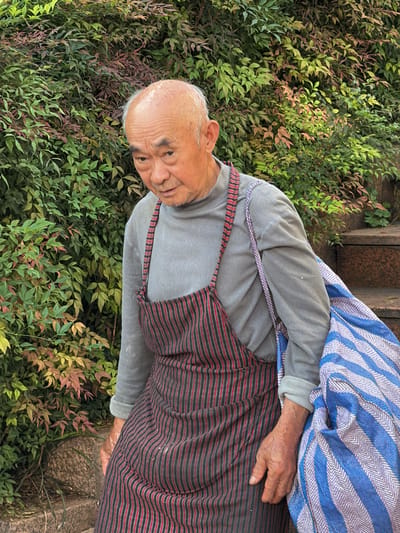 An elderly East Asian man with a bald head and weathered skin is depicted in a medium shot, standing outdoors with a lush green foliage background. He is wearing a grey long-sleeved shirt and a red and black striped apron. A blue and white striped bag is slung over his left shoulder and rests against his right side. The man's head is slightly bowed, and his gaze is directed downwards and to his left. His hands are visible, with his right hand resting on his thigh. The background consists of dense, green leafy plants with some reddish-brown leaves interspersed. To the right of the man, stone steps are visible, suggesting an outdoor, possibly urban, environment. The lighting suggests it is daytime.