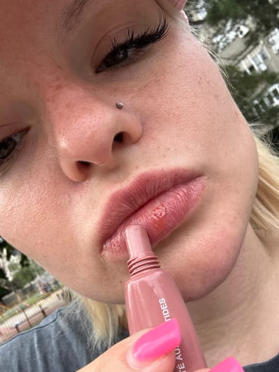 A close-up, outdoor, eye-level medium shot focuses on the lower half of a young Caucasian woman's face. She has fair skin with a scattering of freckles, prominent eyelashes, and a nose piercing. Her lips are painted with a glossy pink balm, and she is holding a matching pink applicator tube. The background is blurred, showing greenery and the facade of a building, suggesting an urban setting. The woman is wearing a dark grey t-shirt, and her blonde hair is visible. The shot appears to be taken in Belgrade, Serbia, based on the context of the location.
