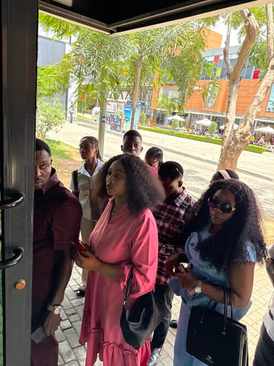 A group of seven people is gathered at the Accra Mall in Itirin, Nigeria. The image is taken through a window, providing a partial view of the environment outside. A man in a maroon shirt is closest to the window, holding a phone. Other people visible include a woman in a pink dress, another in a light blue denim outfit, and others in shirts and casual wear. Some individuals are looking at their phones. The background shows buildings, trees, and pedestrians, typical of a shopping mall environment.