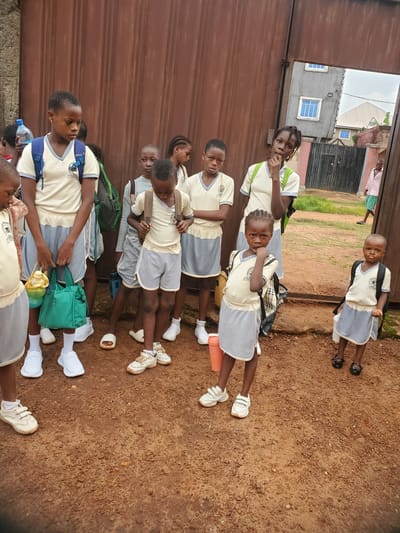 A group of approximately ten children, all wearing similar school uniforms (light yellow shirts and gray shorts or skirts), stand or walk alongside a corrugated metal fence. The setting appears to be in Ukwu-Abwa, Nigeria, based on geographical reference. The children are positioned on a dirt or soil surface, with some carrying backpacks. A few buildings and foliage are visible in the background. Some of the children are looking directly at the camera. One is standing apart from the others on a slight embankment.