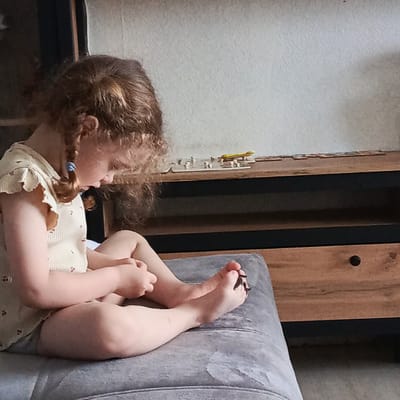 A young child with curly brown hair styled in pigtails sits cross-legged on a grey cushion. The child is wearing a light-colored, patterned top. The child is barefoot, and there appears to be something dark caught between the toes of one foot. The child is looking down at their hands. A wooden cabinet with drawers is visible in the background. The location is unknown, as there is no evidence in the image that it is of Марковский, Russia.