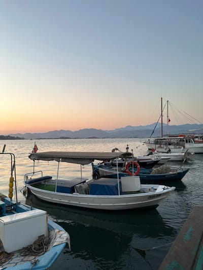 This is a media file of boats docked in a harbor at sunset. The sky is a gradient of soft blues and oranges, indicating it is either sunrise or sunset. The water is calm and reflects the sky, creating a serene atmosphere. Several boats are visible, including a white sailboat with a tall mast and rigging, and smaller motorboats. One motorboat in the foreground is light blue with white trim. The boats appear to be docked for the night. In the background, a mountain range is visible, softened by the distance and haze. The image has a peaceful and tranquil feel. There is some visible text on the boats, though it is difficult to read clearly due to the resolution and angle. On one boat, there appears to be "SU 49" and "1034590". Another boat has "KUMDAL" and numbers below it. A red flag with a white star and crescent, likely the Turkish flag, is visible on one of the boats. This scene is characteristic of a coastal town in Turkey, such as Fethiye, where the mountains meet the sea.