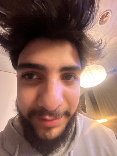 A close-up, angled selfie of a person with dark hair and a beard. The individual's face dominates the frame, showing their eyes, nose, and mouth. A white hooded sweatshirt is partially visible. Overhead, a round light fixture and a circular vent are seen. The image has a slightly warm tone.