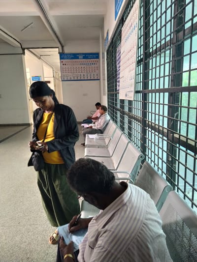 The image shows a waiting area in what appears to be a public space, likely a clinic or government office in Bengaluru, India. Several people are present. A woman in a yellow top and black jacket is looking down at a phone, possibly reading or texting. Another person in a striped shirt is seated and writing on a document. Other individuals are seated further down the row of chairs. The waiting area has metal and plastic chairs. Traffic regulation signs are displayed on the wall. Overhead, there is a grid of metal bars supporting the ceiling, allowing natural light to filter through. The scene suggests people are waiting for services, possibly related to health or government administration.
