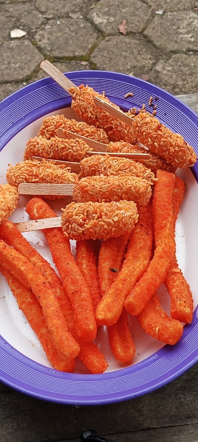 A purple plastic plate sits on a wooden surface. The plate contains an assortment of orange fried food. The food appears to be deep-fried noodles on wooden sticks and some long, thin, orange, deep-fried snacks. The scene is located in the city of Cangkuang, Indonesia at the coordinates -7.065 latitude and 107.551 longitude.