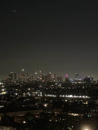 A nighttime aerial view of the Los Angeles skyline is captured, with the downtown area illuminated by countless lights. The buildings vary in height and architectural style, with some displaying colorful illuminated tops, such as purple and pink lights on a few of the towers. In the foreground, the lights of residential areas and lower-rise structures extend towards the viewer, creating a dense urban landscape. Two small, bright, crescent-shaped lights are visible in the dark sky in the upper left quadrant of the image, their nature undetermined. The overall atmosphere is one of a sprawling, vibrant city at night.