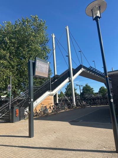 The image depicts the exterior of the Steenwijk train station in the Netherlands. The scene is captured on a sunny day with a clear blue sky.
In the foreground, a paved walkway with a brick-like pattern is visible, casting long shadows from a lamppost on the right. A dark grey lamppost stands prominently on the right side of the image.
To the left, a digital display board shows bus and train departure information. The sign is mounted on a dark grey pole. A tall, leafy tree with green foliage is located behind the display board. Adjacent to the pole is a waste bin with orange detailing on its front. A sign with a circular symbol indicating "ZONE / herhaling" is also visible on a pole to the left of the display.
A pedestrian bridge with a white concrete structure and black railings spans across the scene, leading to a building on the right. The bridge has a modern design, supported by angled white pillars and thin cables. Below the bridge and to the right, a large number of parked bicycles are visible, arranged in racks. Some bicycles are also parked in front of the pedestrian bridge.
The building on the right appears to be part of the train station, constructed with brick. A portion of a train track can be seen under the bridge. The overall impression is a functional and modern urban environment at a transportation hub.