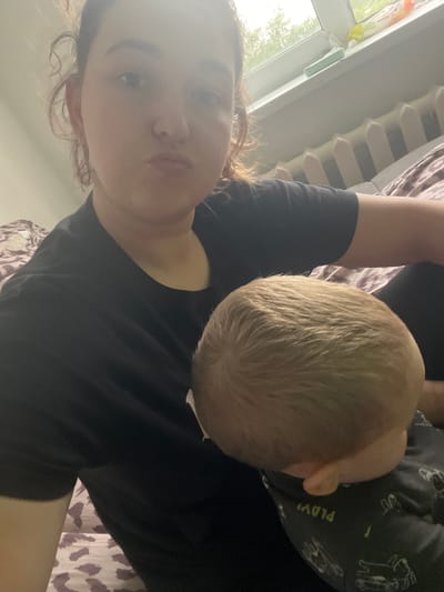 A woman and a baby are lying down, possibly on a bed, in a room. The woman is in the foreground, facing the camera with a neutral expression. She has curly brown hair and is wearing a black t-shirt. Her left arm is visible, bent at the elbow. The baby is nestled beside her, facing away from the camera, with their head visible. The baby has blonde hair and is wearing a dark-colored onesie with a white pattern of animals and text. The background shows a white radiator next to a window, with trees and greenery visible outside. The lighting suggests it is daytime. The woman's clothing and the general atmosphere suggest a relaxed, domestic setting. There is a patterned blanket or duvet on the bed. The text on the baby's onesie is partially visible but difficult to read clearly. The overall scene appears to be a casual, intimate moment between a mother and child.
