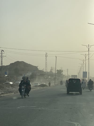 The image captures a dusty street scene in Kandahar, Afghanistan, likely during late afternoon or early morning due to the hazy, warm light filtering through the air. The main subjects are vehicles and people moving along a wide, asphalt road.
In the foreground and midground, several motorcycles are visible. On the left, two individuals ride a motorcycle, both wearing traditional attire. Further back on the right, a black car, possibly a Suzuki WagonR, is prominent, with the letters "NTC" visible in white on its rear window. Other motorcycles and cars are scattered further down the road, and a person is seen walking on the roadside on the left.
The setting is characterized by a dry, arid environment. Mountains or large hills form the backdrop, with some structures and utility poles/towers discernible against their slopes. Power lines crisscross the hazy sky, connecting numerous utility poles and at least two distinct cell towers. The air appears thick with dust, creating a soft, diffused light and reducing visibility in the distance. Piles of dirt or construction debris are visible along the sides of the road. Streetlights with multiple arms line the right side of the road, extending into the background. A billboard with indistinct text is also visible on the right.
The overall scene depicts a common daily activity of transportation in a semi-urban or rural area, marked by the distinctive regional characteristics of traditional dress, prominent motorcycle usage, and a dusty, arid landscape.