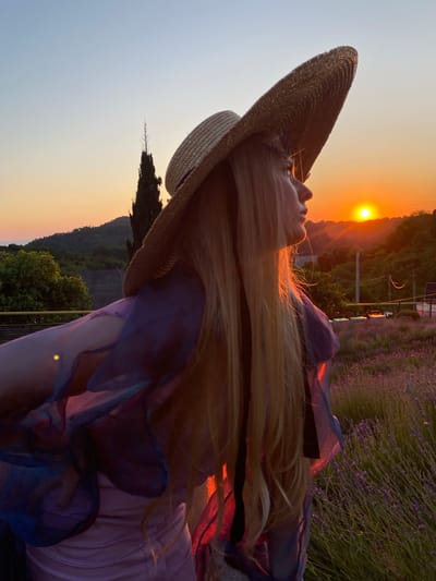An outdoor medium shot shows a woman with long blonde hair wearing a wide-brimmed straw hat and a flowing purple and pink dress in the city of Verkhneye Uchdere, Russia. The woman is viewed from the side, looking towards the right of the frame at a sunset. The sun is a bright orange orb low in the sky, casting a warm glow on the scene. She is standing in a field of lavender flowers, with rolling hills and trees in the background. The sky is clear and transitions from a soft blue at the top to a warm orange near the horizon. The overall mood is serene and peaceful, captured during the golden hour.