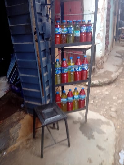 The image depicts an outdoor stall in Kabong, Nigeria, where various liquids are displayed in plastic bottles on a metal shelving unit. The stall is situated next to a brick building with a weathered facade. A dark, blue metal structure, possibly a staircase or part of a larger construction, is visible on the left side of the frame.
The shelving unit holds multiple rows of bottles filled with liquids of different colors, predominantly red and yellow, with some appearing clear or green. The bottles are arranged neatly, suggesting they are for sale. A small, dark stool or low table with a decorative top is placed in front of the stall. The ground around the stall appears to be uneven and dusty, with some damp patches.
There is no visible indication of the time of day or weather, though the lighting suggests it could be daytime. The scene appears to be a typical street-side vendor setup. No people are clearly visible in the frame, and no specific emotions are conveyed. The focus is on the products and the stall itself. Some text might be present on the bottle labels, but it is not legible in the provided image. The overall impression is that of a small business operating in a possibly busy, outdoor urban environment.
