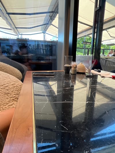 The image shows a close-up view of a table at an outdoor cafe in Baku, Azerbaijan. The table has a black marble top with a wooden edge and a brass trim. On the table are various items, including a clear glass, a dark-colored pot, a salt and pepper shaker set, a sugar dispenser, and a cell phone. In the background, through a glass partition, can be seen patrons sitting at other tables, umbrellas providing shade, and lush green trees. The overall setting suggests a pleasant and relaxed dining experience in a well-appointed outdoor seating area.