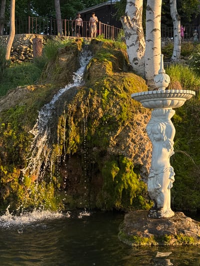 The image captures a scene in Krasny Klyuch, Russia. A moss-covered rocky formation with a small waterfall is prominent. A classical-style sculpture rests atop the rock. Water flows from the sculpture into a dark pool. A man reclines on a platform supported by birch trees and a metal railing is visible in the background. A wooden staircase is near the platform.