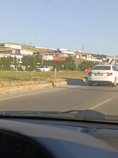 A white car with license plate "34 DAB 462" is driving on a road in Yahşihan, Turkey. In the background, there are industrial buildings and a hill under a clear blue sky, suggesting it is daytime. Another red car is visible further down the road. The scene appears to be a semi-urban or industrial area. The perspective is from inside a vehicle, looking out through the windshield. The weather is clear and sunny.