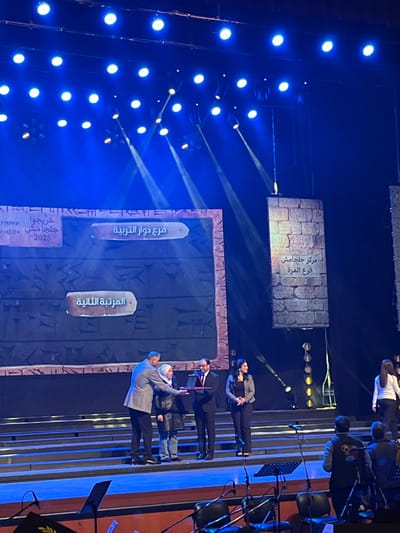 The scene depicts an awards or recognition ceremony taking place on a brightly lit stage within the city of Al-Qanawat Municipality, Syria. The stage features multi-tiered steps and a large backdrop resembling ancient stone tablets or cuneiform, displaying Arabic text. The text on the left reads "خريجوا جلجامش 2025" (Graduates of Gilgamesh 2025), with "فرع دوار التربية" (Education Roundabout Branch) and "المرتبة الثانية" (Second Place) displayed centrally. On the right, a vertical banner reads "مركز جلجامش فرع المزة" (Gilgamesh Center Al-Mezzeh Branch).

In the center of the stage, four individuals are engaged in an interaction. A man in a grey suit jacket stands on the left, extending his right hand to shake the hand of a woman in a white hijab, black jacket, and blue jeans, who stands facing the audience's left. To her right, a man in a dark suit and glasses holds a red-cased object, possibly an award or plaque, presenting it. A woman in a black blazer and dark pants stands observing to the right of the presenter.

The stage is illuminated by numerous overhead blue lights and spotlights. In the foreground, an orchestra pit or performance area is visible, with multiple microphones on stands, hinting at musical accompaniment. Individuals are partially visible in this area, including one person wearing a dark jacket with a yellow emblem. On the far right of the stage, another woman in a white shirt and dark pants is descending the steps.