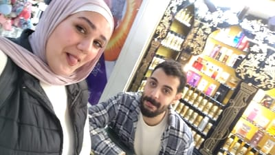 The image portrays two individuals inside what appears to be a retail establishment in Barza Municipality, Syria. A female subject occupies the left foreground, facing the camera with a light complexion, dark eyebrows, and visible eyeliner. She wears a light pink or beige hijab, a white long-sleeved top, and a black quilted vest, displaying a smile with her tongue extended. Behind her, on the far left, a blurred display of colorful hair accessories and a partially visible purple object with text are discernible. To the right and slightly behind the female, a male subject is visible, also looking towards the camera with his tongue extended. He has dark hair and a dark beard, wearing a white t-shirt under a blue and white plaid flannel shirt. The background on the right consists of multiple ornate display shelves, dark in color with prominent gold-colored classical and floral patterns. These shelves are filled with numerous bottles, consistent with perfumes or colognes, varying in color and design, illuminated by bright interior lighting.