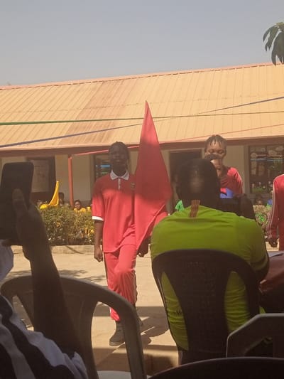 The image depicts an outdoor daytime scene in Narayi, Nigeria, likely at a school or community event. In the midground, a young female, approximately in her teens, walks towards the right while looking forward. She wears a red polo shirt with a white collar and cuffs, red pants, and dark-colored shoes. She holds a large, triangular red flag vertically with her right hand, positioned to her left. Her hair is styled in dark braids. Partially visible to her right and slightly behind her is another individual, also appearing to be a young female in a red top with braided hair. Further to the right, a third individual in a red and white uniform is partially visible.
In the immediate foreground, to the left, a person's left arm and hand are visible, holding a black smartphone upright, alongside the top portion of a dark-colored plastic chair. To the right, the back of a person wearing a bright lime green short-sleeved shirt with dark stripes on the collar/shoulder area is visible, seated in a dark-colored plastic chair.
The background features a single-story building with a light brown corrugated metal roof. Colorful streamers in red, blue, and green are strung across the scene in front of the building. Below the streamers and in front of the building, a line of green bushes partially obscures additional individuals, some wearing yellow tops, who appear to be observing the event. The ground surface is light-colored and unpaved. The sky is clear and bright blue, indicating clear weather.