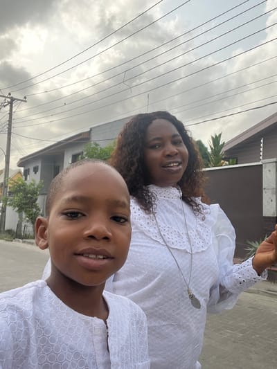 A photograph taken in Iranla, Nigeria, depicts an adult female and a child posing outdoors on a paved surface under an overcast sky. In the foreground, slightly to the left, a child with light brown skin wears a white, short-sleeved top with a circular textured pattern, looking directly at the camera with a slight smile. Behind and to the right, an adult female with dark, curly hair and light brown skin is visible from the chest up, smiling with her mouth slightly open, looking slightly off-camera to the right. She wears a white high-necked blouse featuring intricate eyelet lace details and ruffles on the collar and cuffs. A long, silver-colored necklace with an oblong, decorative pendant hangs around her neck. Her left hand, with fingers slightly splayed, is partially visible on the right side of the frame. The background features residential structures: a light-colored building with dark windows and a peaked roof is visible on the left, while a darker wall or building section topped with a security fence (electric fence insulators/wires) is present on the right. Green foliage is interspersed between the subjects and background buildings. A utility pole with multiple overhead wires is visible on the far left. The ground is a paved surface, consistent with a road or sidewalk.