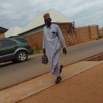 A man wearing a light blue traditional robe and a patterned cap is walking on the side of a road. He is carrying a black and white patterned bag in his left hand. A dark green SUV is parked on the road to his left. In the background, there are buildings with white roofs and brick walls, some of which are under construction with scaffolding and ladders. The sky is overcast, suggesting it is either early morning or late afternoon, and the weather is cloudy. The scene appears to be in an urban or semi-urban area in Gombe, Nigeria. There is some text visible on the brick wall in the background, but it is not clearly legible.
