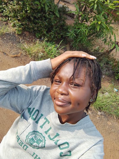 A young Nigerian woman is captured in a selfie-style shot, smiling directly at the camera. She has dark skin, braided hair, and a prominent dimple on her left cheek. Her left hand is raised, resting on the top of her head, possibly shielding her eyes from the sun. She wears long, light-colored acrylic nails.
She is dressed in a light grey long-sleeved sweatshirt with green text and a circular crest logo. Due to the selfie angle, the text on her sweatshirt is reversed. When viewed correctly, visible parts of the text appear to say "COLLECTION" (or "COLLEGION"), "DEPT" (likely "DEPARTMENT"), and within the crest, "STATE UNIVERSITY" and "FOUNDED". She also wears a small, dark clover-shaped earring in her right ear.
The setting is outdoors in Nsukka, Nigeria, under bright daylight conditions, suggesting a sunny day. The background consists of abundant green foliage, including bushes, grass, and small trees, growing on reddish-brown soil or dirt. The ground appears unpaved, indicating a natural or rural environment. The woman's expression is joyful and relaxed.