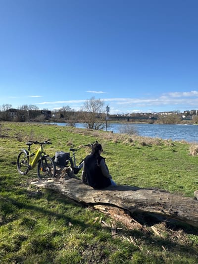 The image depicts a sunny outdoor scene on the grassy bank of the Ruhr River in Hattingen, Germany. In the foreground, a single individual is seated facing away from the viewer, resting on a large, weathered, horizontal log. The person wears a dark vest over a lighter long-sleeved top and light blue jeans, with dark hair pulled back. Two bicycles are parked on the grass to the left of the log; the closer one is a neon yellow/lime green electric mountain bike, and the other is a dark-colored touring or e-bike equipped with a dark rear pannier bag.

The midground features the wide, blue-grey Ruhr River flowing horizontally across the frame. On the opposite bank, sparse, mostly bare deciduous trees and some distant buildings are visible. A white square sign on a pole is partially visible among the vegetation on the riverbank to the right. In the far midground, a multi-span bridge crosses the river. Beyond the bridge and river, the background reveals a town, consistent with Hattingen, situated on a rising hillside with numerous buildings and trees. The sky is clear blue with a few wispy white clouds, indicating bright daylight conditions.
