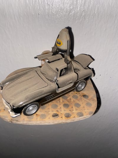 This media file features a dusty, dark-colored toy car placed on a piece of woven material. The car appears to be a vintage model, with its doors slightly ajar. A portion of its front headlight and bumper are visible, with a yellow accent on the headlight. The woven material is light brown and has a crisscross pattern, with some darker circular areas visible within the weave. The background consists of a textured, off-white surface on the left and a dark grey surface on the right, with a shadow cast by the car and woven material. The scene appears to be indoors, possibly in a storage area or a room that has not been recently cleaned, given the layer of dust on the toy car. The lighting suggests it might be daytime, but artificial light could also be the source. There is no visible text in the image.