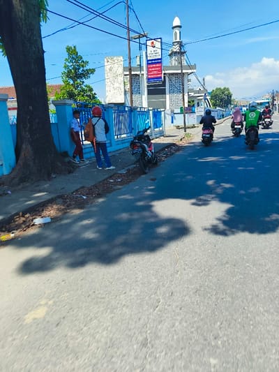 The image shows a street scene in Margahayu, Indonesia, at the coordinates -6.982 latitude and 107.563 longitude. The scene is in front of a building with a blue and white fence and a mosque behind it. The street is paved with asphalt. Three men on motorcycles are riding towards the camera, and two boys are standing near the fence. The boys are wearing school uniforms. The street is mostly empty, except for the motorcycles and the two boys. The sky is blue with a few clouds. The sun is shining, casting long shadows from the building and the people. A large tree stands on the left side of the image, creating a shadow across the sidewalk and street. There is debris on the sidewalk, including a paper bag and a discarded branch. The building behind the fence appears to be some type of business. A sign on the building has the text "A. RM. HARI - BAKSO" on it.