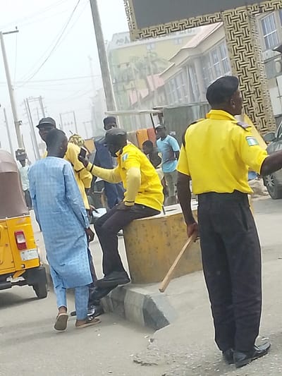 The image captures an outdoor urban street scene in Gandu Albasa, Nigeria, under a hazy sky. In the immediate foreground, a male individual wearing a yellow short-sleeved uniform shirt with a Nigerian flag patch on the left arm, dark trousers, and a black beret stands with his back largely to the viewer, holding a wooden stick in his right hand, extending downwards. To his left, another male, similarly dressed in a yellow uniform shirt, dark trousers, and a black beret, is seated on a large, rectangular yellow concrete barrier or curb block. This seated individual holds a smartphone in his right hand, oriented towards a male in a light blue patterned long tunic (agbada) who stands facing away from the camera. A yellow three-wheeled auto-rickshaw is partially visible on the far left. Multiple other male individuals in civilian attire are present in the mid-ground and background, some appearing to be standing or conversing. The background reveals various urban structures, including a multi-story building with a gold-colored facade and a visible "AO^" logo, and another building featuring a decorative geometric patterned exterior. Overhead utility poles and electrical lines crisscross the pale sky. The asphalt road surface exhibits minor wear, and a concrete curb line is visible.