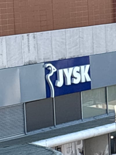 The image shows a sign for "JYSK" on the exterior of a building. The sign is illuminated with white lettering on a dark blue background. Above the JYSK sign, there is a light-colored textured wall, and above that, a brown brick wall with a grid pattern. Below the sign, there are windows and a gray section of the building. The scene is consistent with the JYSK store located at **24-25 St. Patrick's Street, Cork, Ireland**.