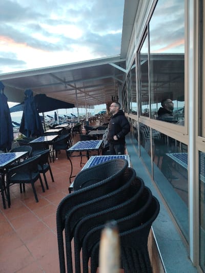 An outdoor cafe terrace in a coastal area of Tanger, Morocco, is depicted at either dusk or dawn under an overcast sky with hints of soft orange and pink. The terrace features numerous black wicker-style chairs and square tables with blue and white mosaic-patterned tops. Some chairs are stacked to the right foreground. A white canopy extends over the terrace. To the right, a building facade with large reflective glass windows runs along the length of the terrace.
A sole male individual is visible, standing midway down the terrace, facing left and looking towards the distant horizon or sea. He wears a dark hooded top and dark trousers, has a beard, and wears glasses. His reflection is clearly visible in the adjacent glass windows. In the background to the left, the sea is visible, alongside several large, folded dark beach umbrellas and more cafe seating. A blurred, lit cigarette or hand-rolled cigarette, exhibiting a glowing ember, is prominently centered in the immediate foreground, suggesting it is held by the person taking the photograph.