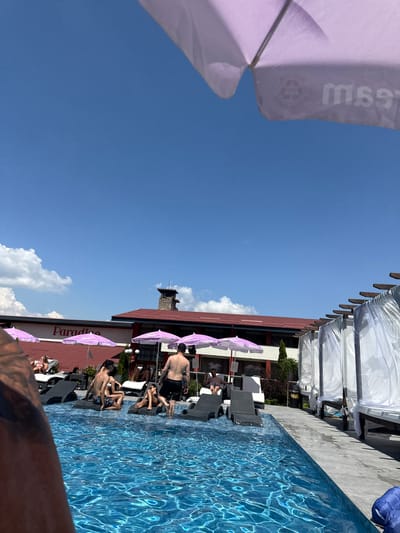 A group of people are enjoying a sunny day by a swimming pool at a resort. The scene is outdoors, with a clear blue sky and scattered white clouds. The swimming pool is filled with bright blue water, reflecting the sunlight. Several lounge chairs are set up around the pool, some occupied by people relaxing.
Purple umbrellas provide shade, with some displaying text that reads "Ice Cream." A building with a red roof is visible in the background, featuring multiple levels and windows. Several white canopies are set up on a raised platform above the lounge chairs.
Some individuals are in swimwear, with one person standing and another sitting in a lounge chair. Other people are seen lounging on chairs near the pool. The overall atmosphere appears relaxed and leisurely, suggesting a vacation or a day of leisure. The time of day is likely midday, given the bright sunlight and clear sky. The location cues, such as the building architecture and the presence of a pool and lounge area, suggest a resort or hotel environment.