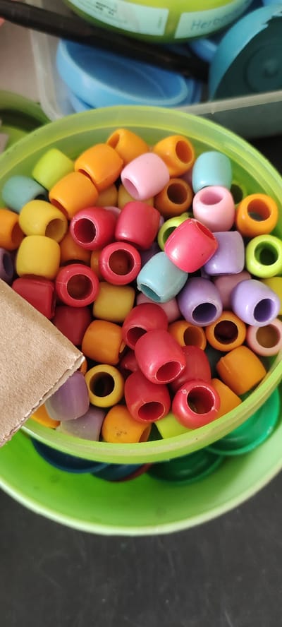 A green plastic container filled with colorful plastic beads. The beads are primarily red, yellow, orange, green, blue, and purple. There is a small piece of cardboard sticking out of the container. The container sits on a dark surface, likely a table or countertop. The image was taken in the city of Sidoarjo, Indonesia, at the coordinates -7.443, 112.71.